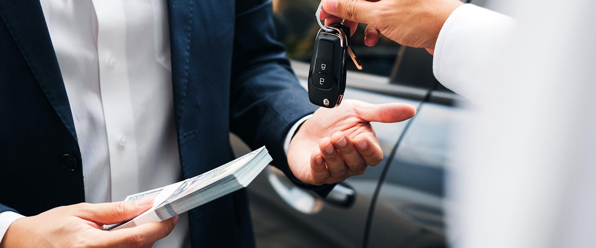 A person hands over car keys while another holds a stack of cash, suggesting a transaction, in front of a car.